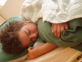 A boy with dark hair is sleeping on his side in a bed with green sheets and pillow